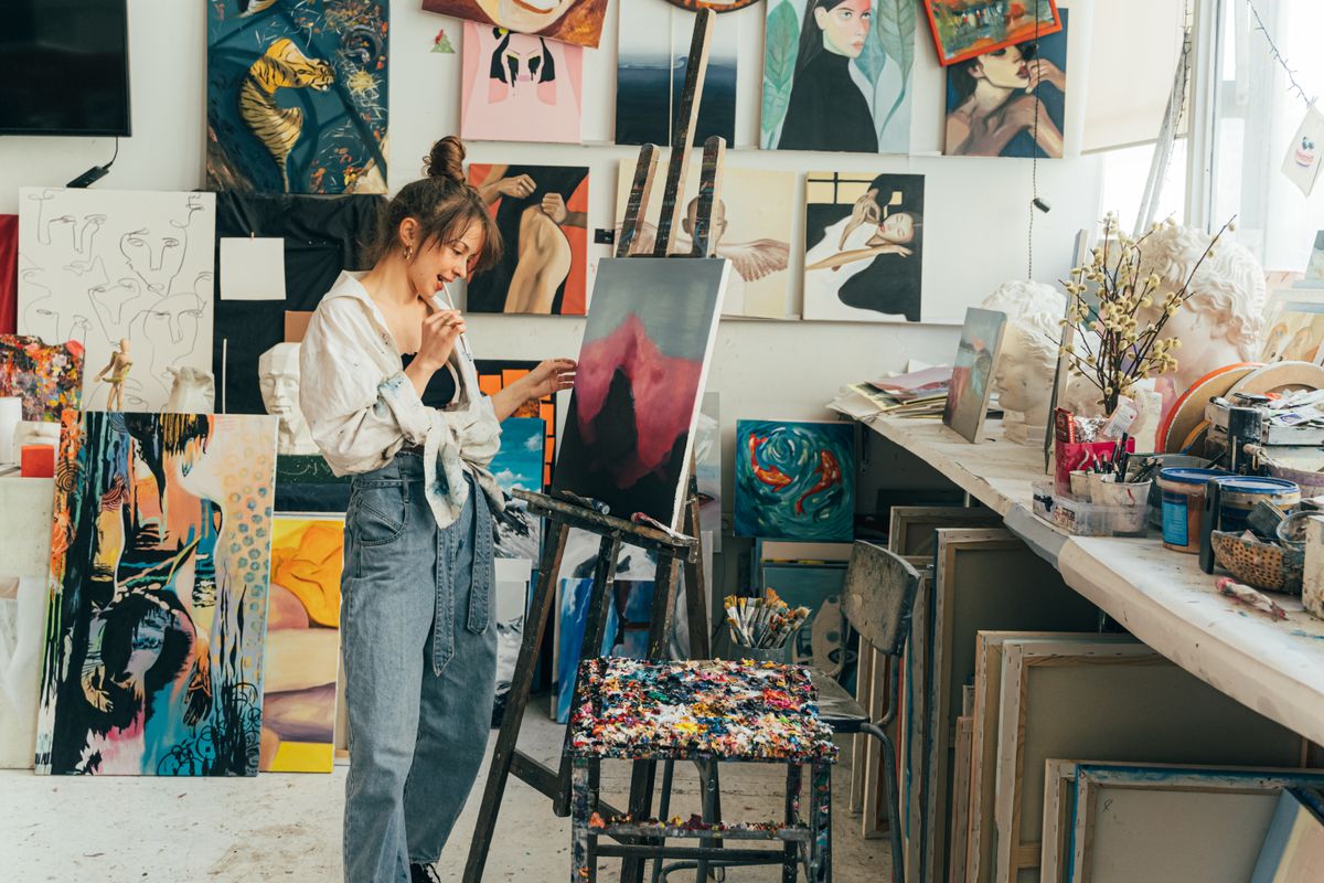 An artist painting at an easel in a studio hung with finished canvases